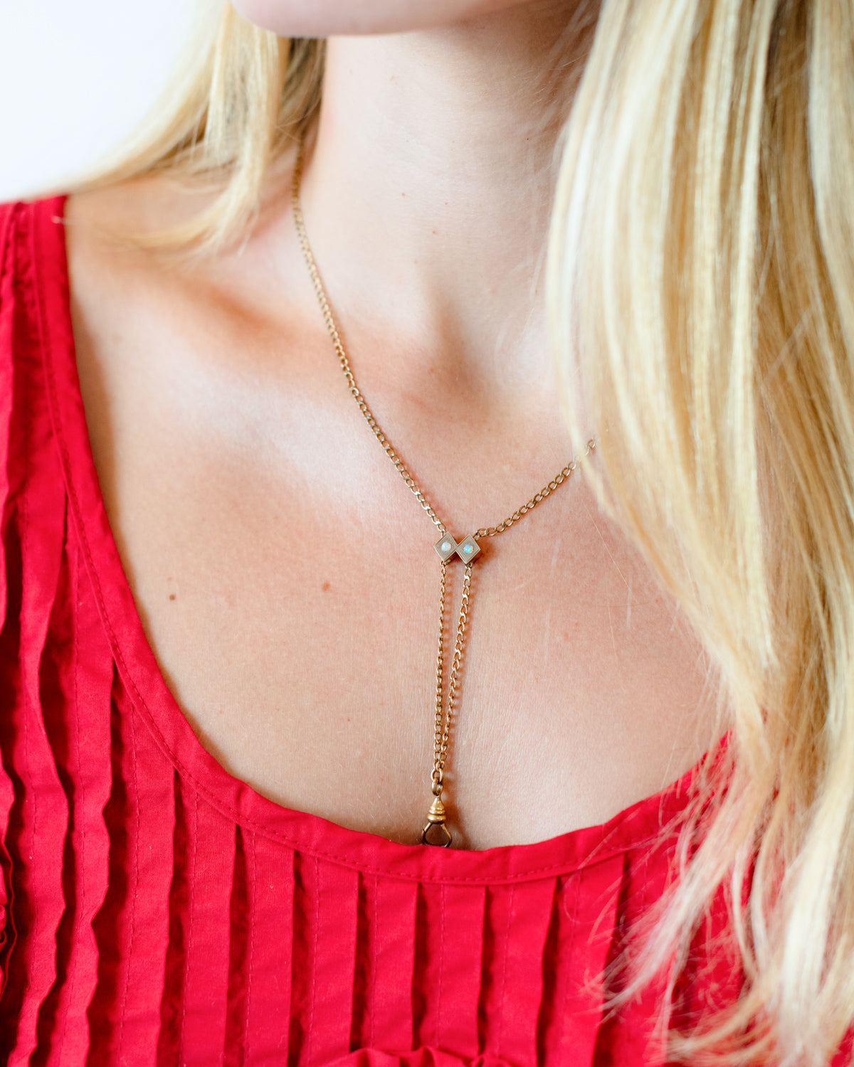 A close-up of a model wearing a delicate gold lariat necklace with a small bow-shaped charm with opal stones and a working dog clip, styled with a red top.