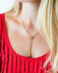 A close-up of a model wearing a delicate gold lariat necklace with a small bow-shaped charm with opal stones and a working dog clip, styled with a red top.