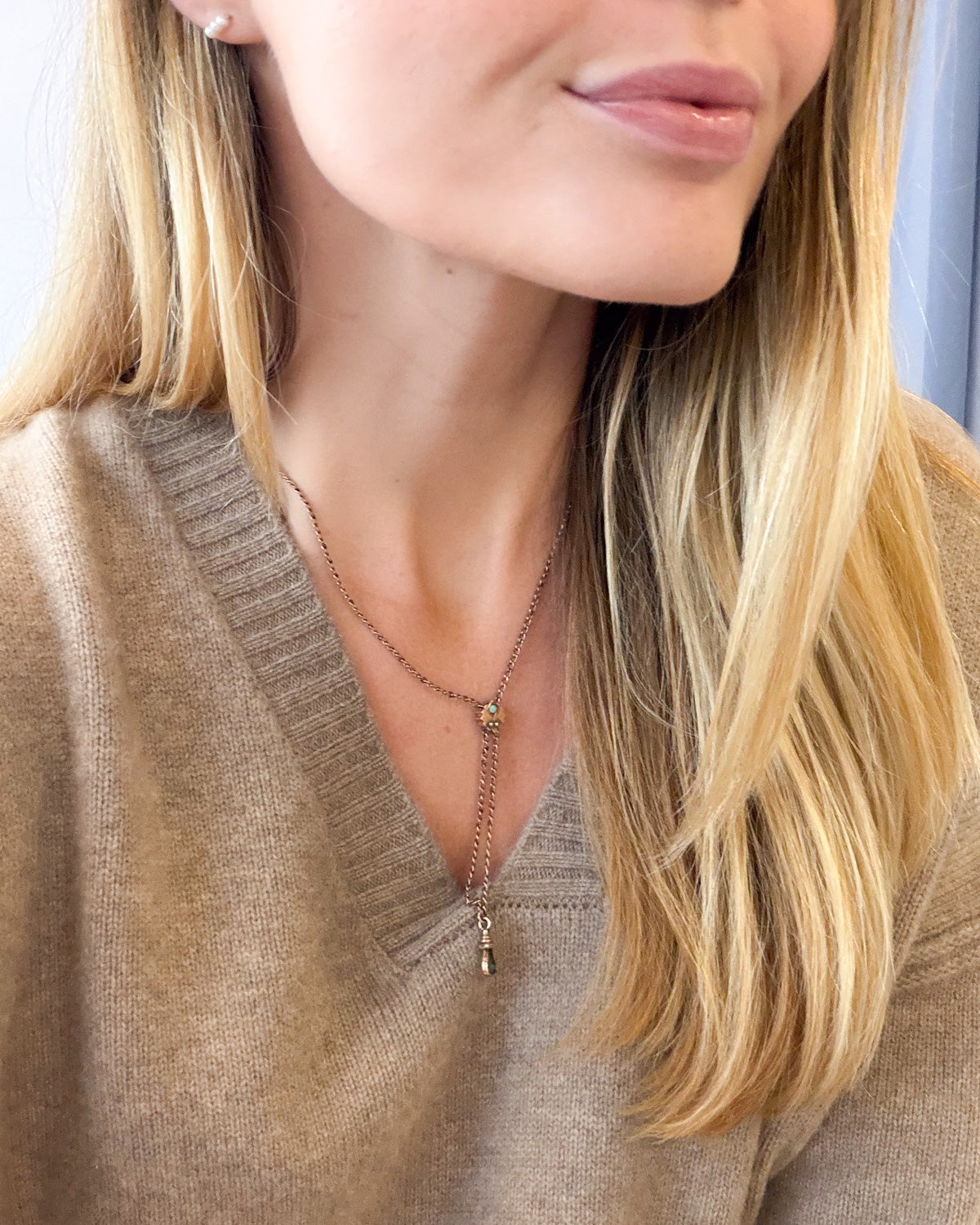 A close-up lifestyle shot of a woman wearing a delicate gold lariat necklace with a small bead and drop pendant, styled with a beige knit V-neck sweater.