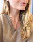A close-up lifestyle shot of a woman wearing a delicate gold lariat necklace with a small bead and drop pendant, styled with a beige knit V-neck sweater.