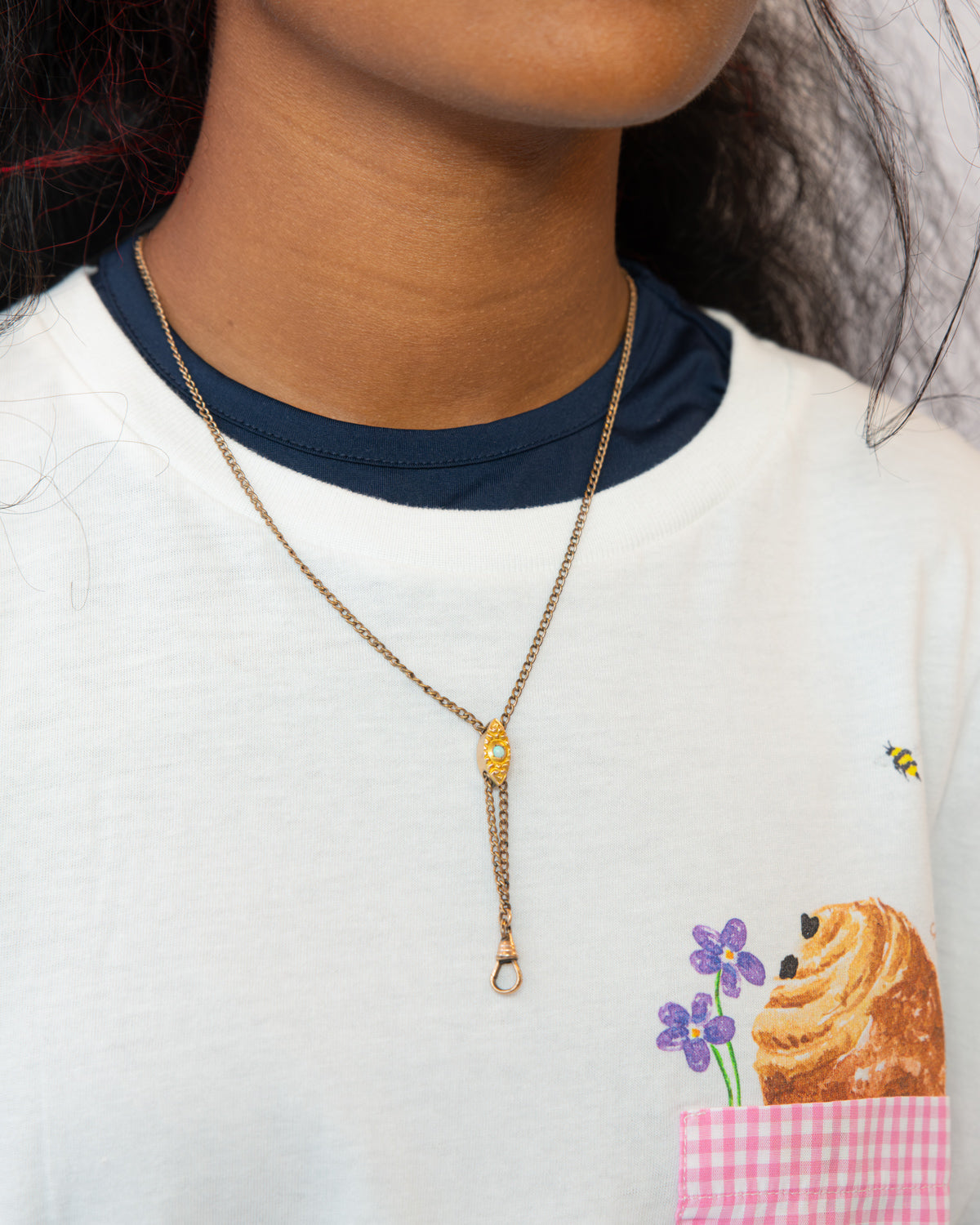 A close-up of a person wearing a delicate gold Victorian-era chatelaine slider necklace with a small gold charm and drop ring, styled over a white ringer T-shirt with a navy collar and a cartoon pastry pocket.