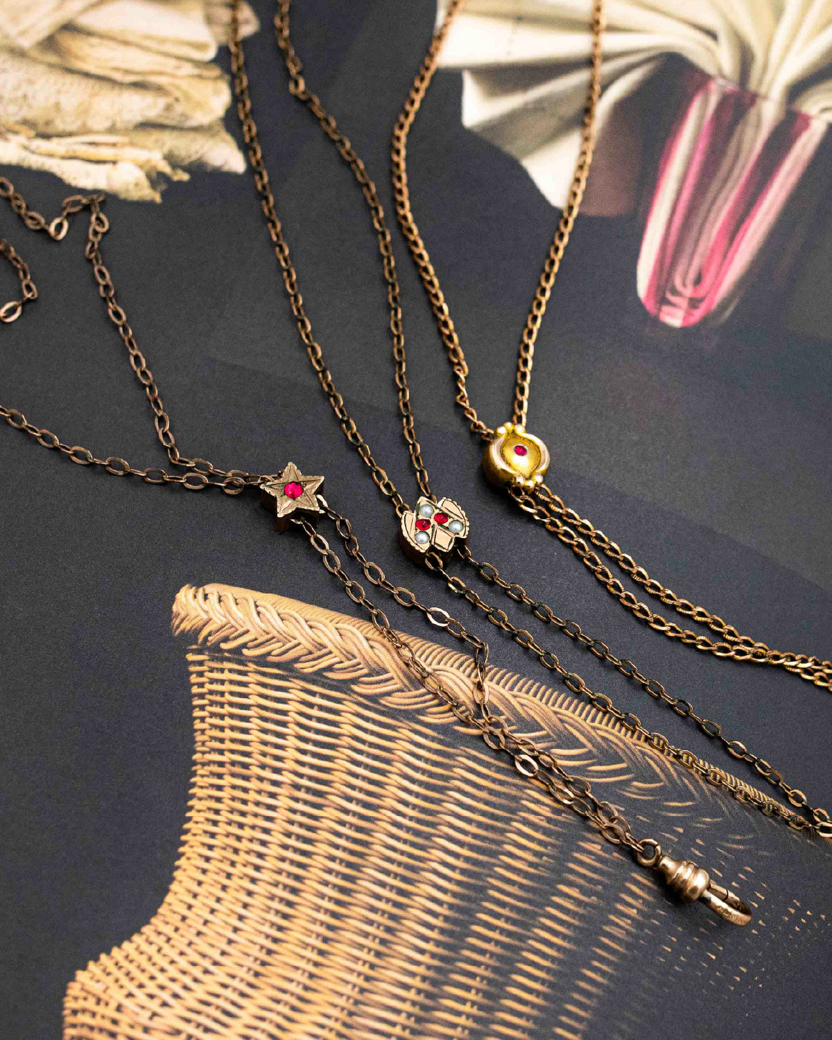 Three antique Victorian-era lariat chatelaine slider necklaces with decorative pendants arranged on a dark textured surface with a woven basket and dried flowers as props. The necklaces feature a star pendant with a red gem, a floral pendant with a red gem and clear stones, and a small gold heart pendant.