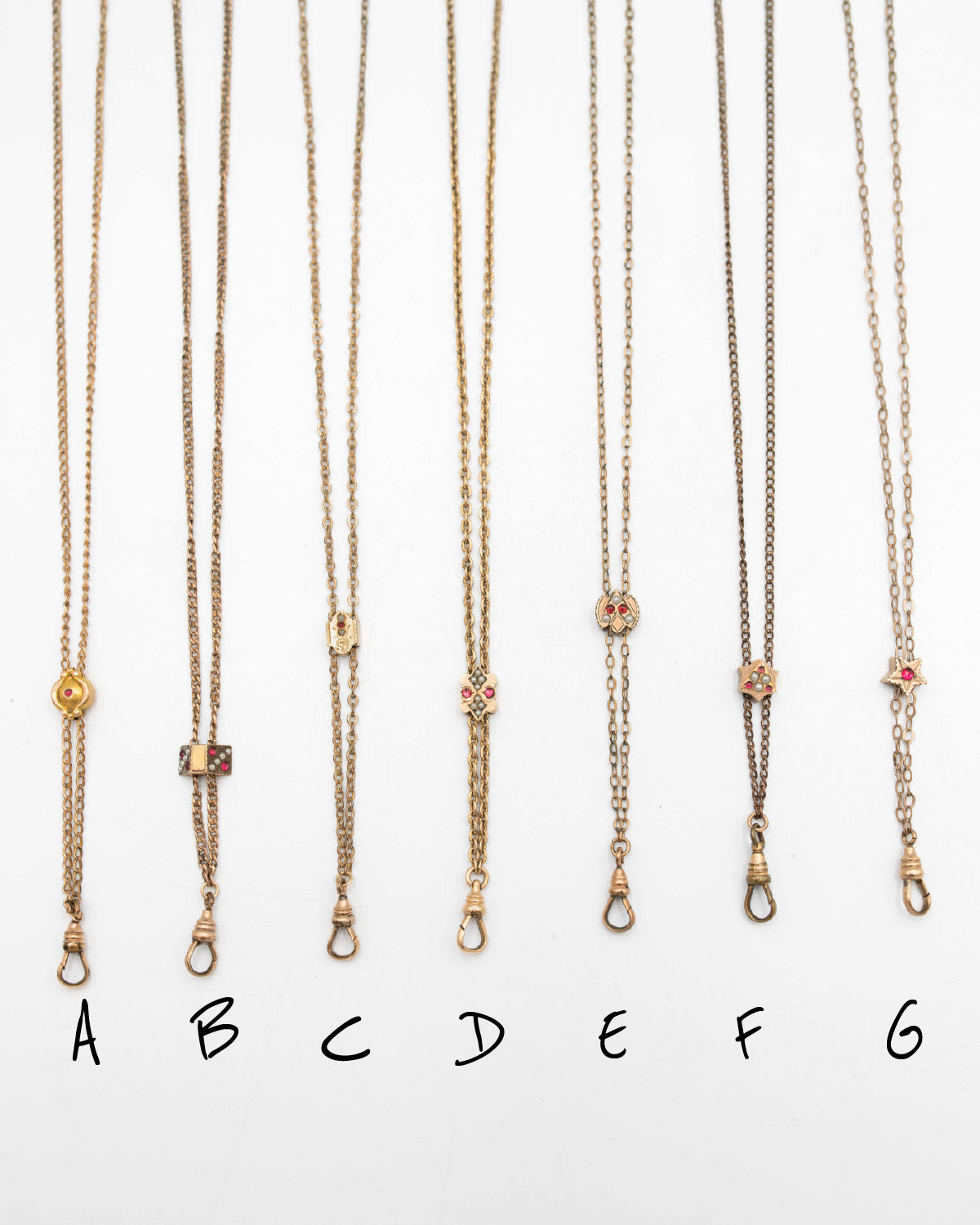 Six gold-filled and solid gold Victorian-era chatelaine lariat slider necklaces with small pendant charms are displayed vertically on a white background, labeled A through G below each.