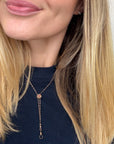 A close-up of a woman wearing a delicate rose gold lariat necklace with a small round bead and a dangling loop, paired with a dark navy top.