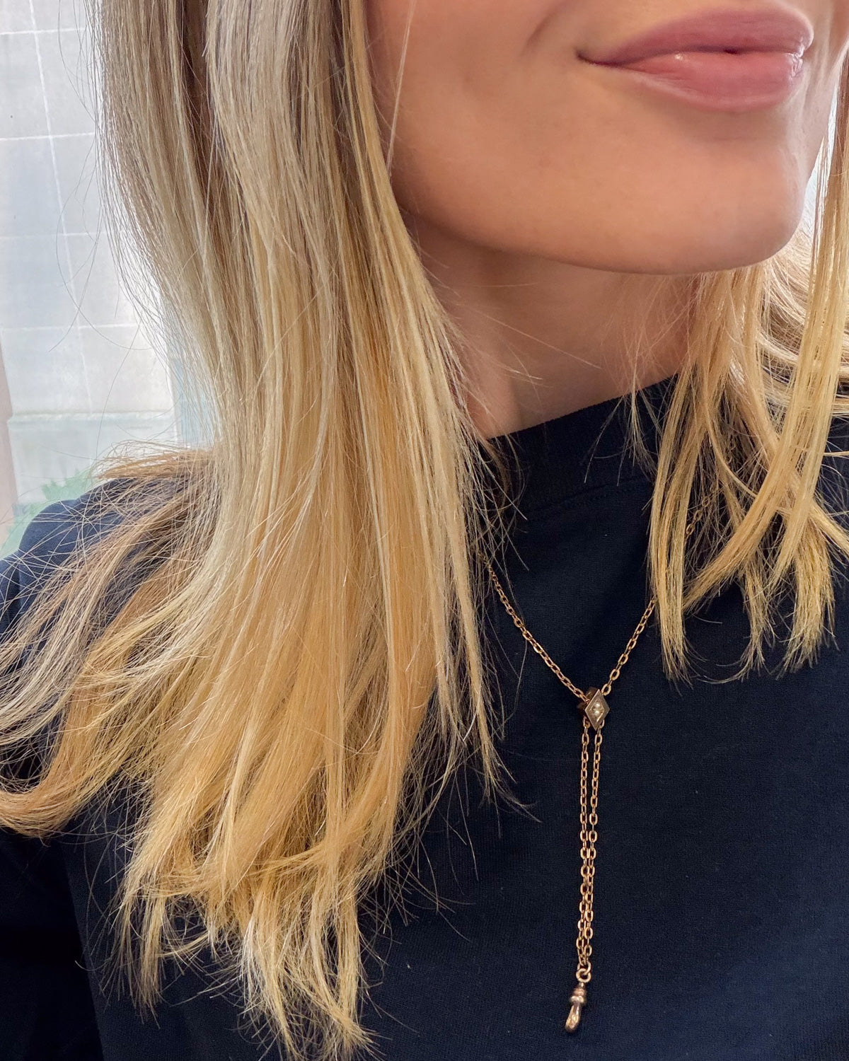 A close-up of a woman wearing a delicate gold lariat chatelaine necklace with a long drop chain and a small dangling charm, paired with a black top.