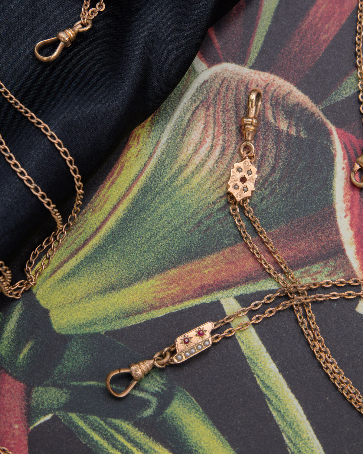 Gold-tone chain jewelry with a small rectangular charm featuring three red glass ruby stones and a row of seed pearl rhinestones, laid on a dark satin fabric over a tropical floral print background.