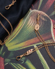 Gold-tone chain jewelry with a small rectangular charm featuring three red glass ruby stones and a row of seed pearl rhinestones, laid on a dark satin fabric over a tropical floral print background.