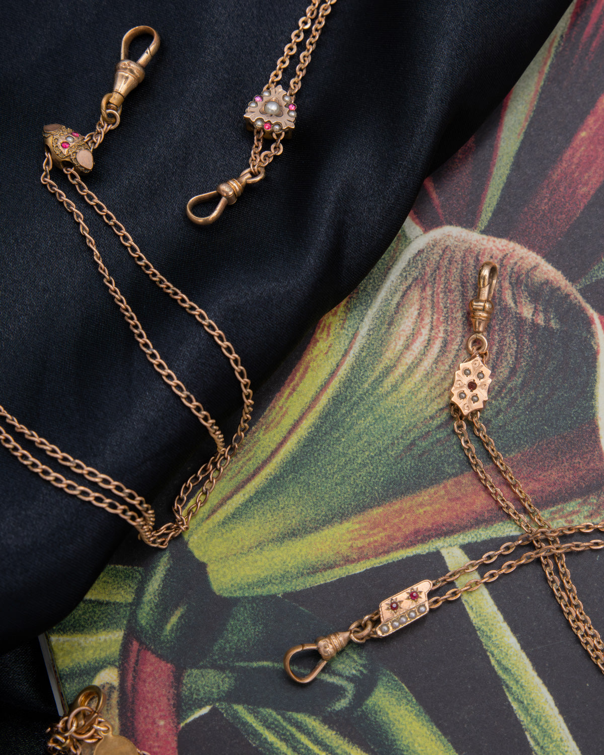 Gold-filled Victorian-era chatelaine necklaces with dog clip clasp connectors and small floral charms, arranged on a dark navy fabric and a tropical leaf print surface.