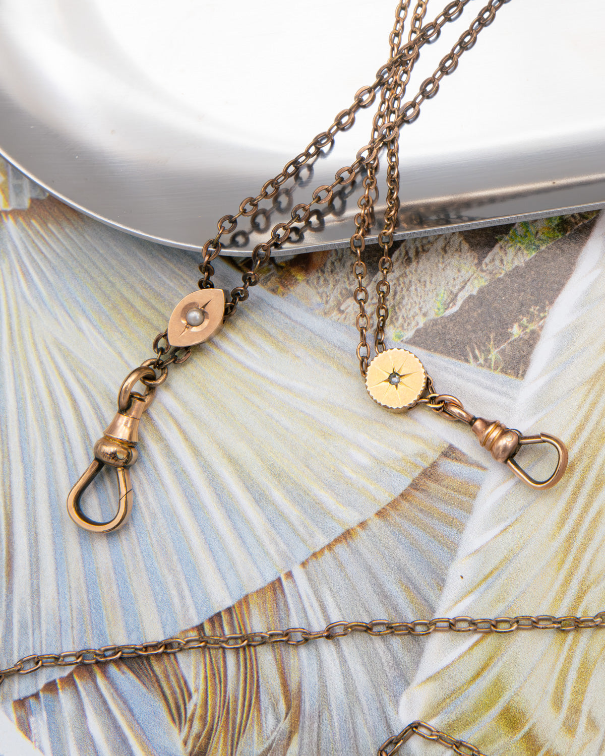 Two antique bronze chain chatelaine necklaces with round disc charms and lobster clasp closures, displayed on a textured beige and blue leaf-patterned surface.