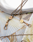 Two antique bronze chain chatelaine necklaces with round disc charms and lobster clasp closures, displayed on a textured beige and blue leaf-patterned surface.