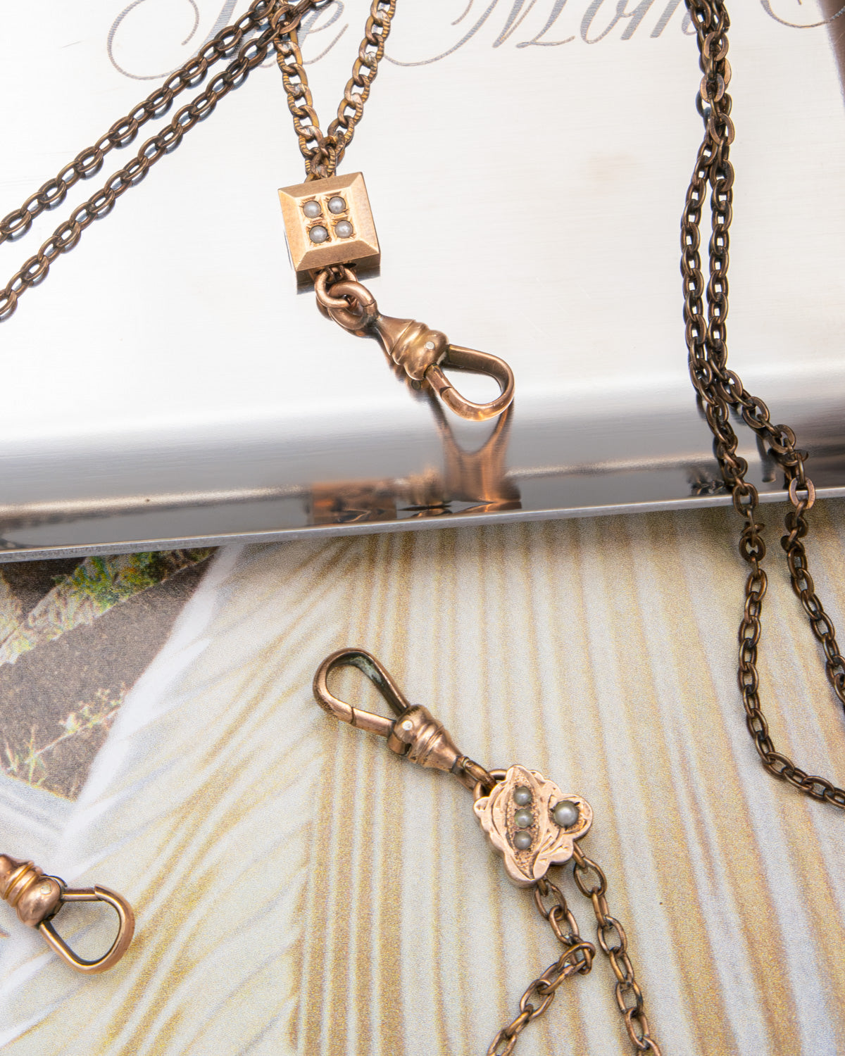 Close-up of antique bronze-toned gold-filled chatelaine chain necklaces with decorative charms, including a small square pendant with four inset stones and a floral charm with three round stones, arranged on a light surface.