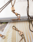 Close-up of antique bronze-toned gold-filled chatelaine chain necklaces with decorative charms, including a small square pendant with four inset stones and a floral charm with three round stones, arranged on a light surface.