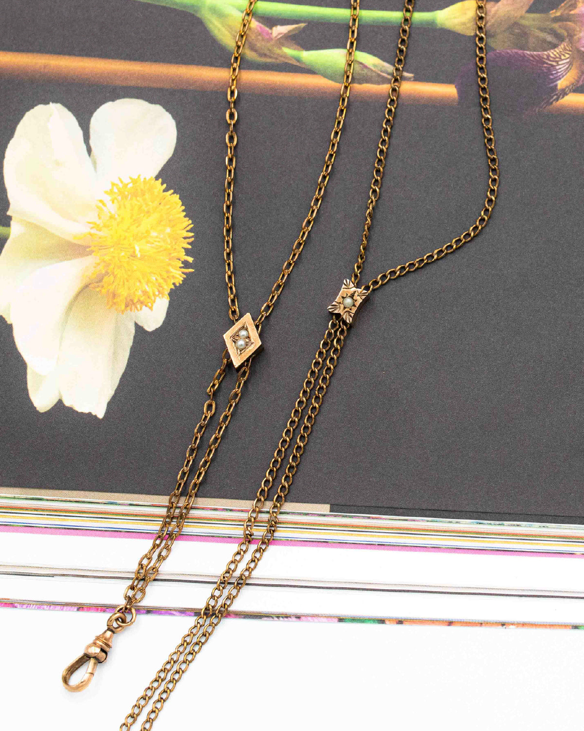 An antique bronze double-strand Victorian-era gold-filled chatelaine necklace with a small diamond-shaped crystal pendant and a larger square crystal pendant, displayed on a dark book cover next to a white flower.