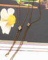 An antique bronze double-strand Victorian-era gold-filled chatelaine necklace with a small diamond-shaped crystal pendant and a larger square crystal pendant, displayed on a dark book cover next to a white flower.