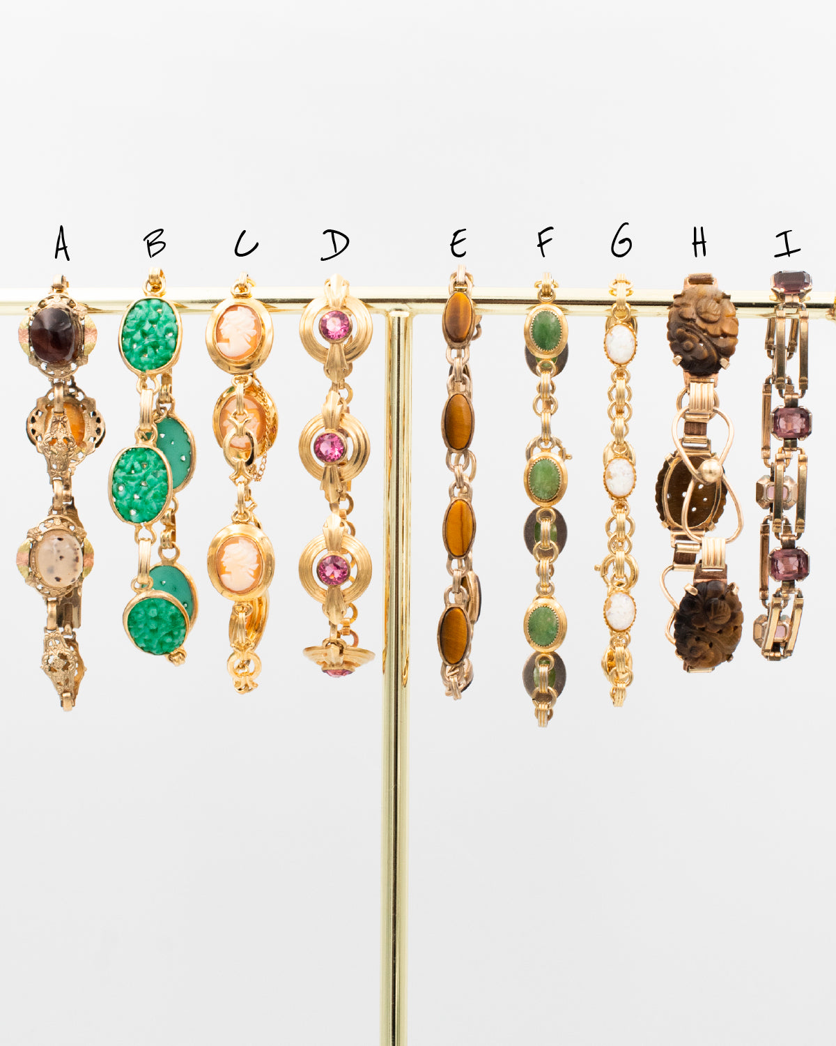 Collection of vintage gold-filled bracelets from the 1930s–1960s featuring cameos, carved stones, Peking glass, and gemstone cabochons displayed on a gold stand