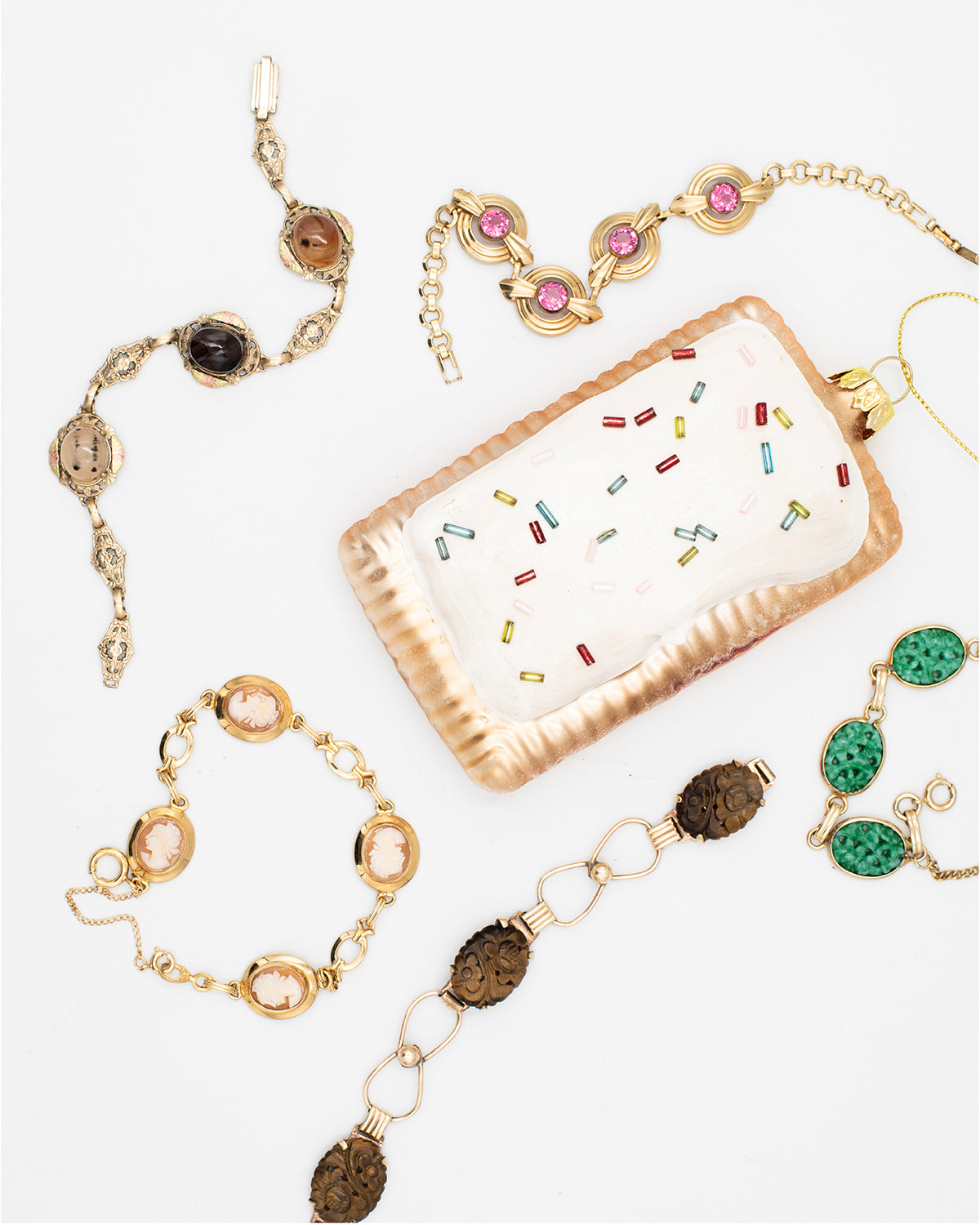 Five gold-fill vintage bracelets with cameos, carved peking glass, carved tiger's eye, pink glass and agate on a white background around a pop-tart holiday ornament