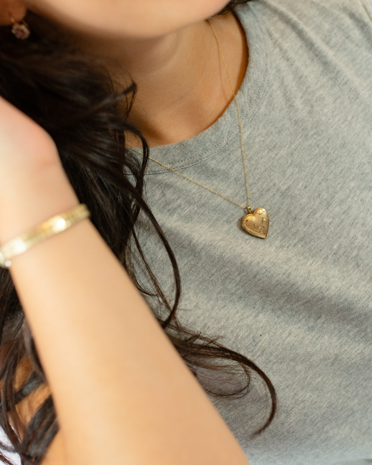 A close-up lifestyle shot of a person wearing a delicate gold heart locket pendant necklace on a fine chain, paired with a gray heathered t-shirt and a gold bangle bracelet.