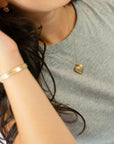 A close-up lifestyle shot of a person wearing a delicate gold heart locket pendant necklace on a fine chain, paired with a gray heathered t-shirt and a gold bangle bracelet.