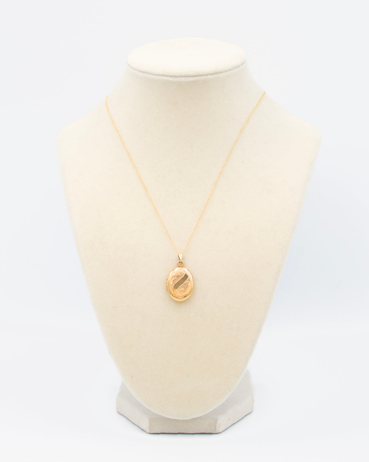 Vintage oval gold locket with unique engraving displayed on a white jewelry bust