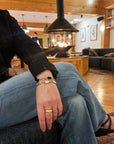 A person sitting in a cozy wood-paneled room wearing a stack of vintage onxy and gold-fill bracelets on one wrist, with blue jeans and a black lace top visible.
