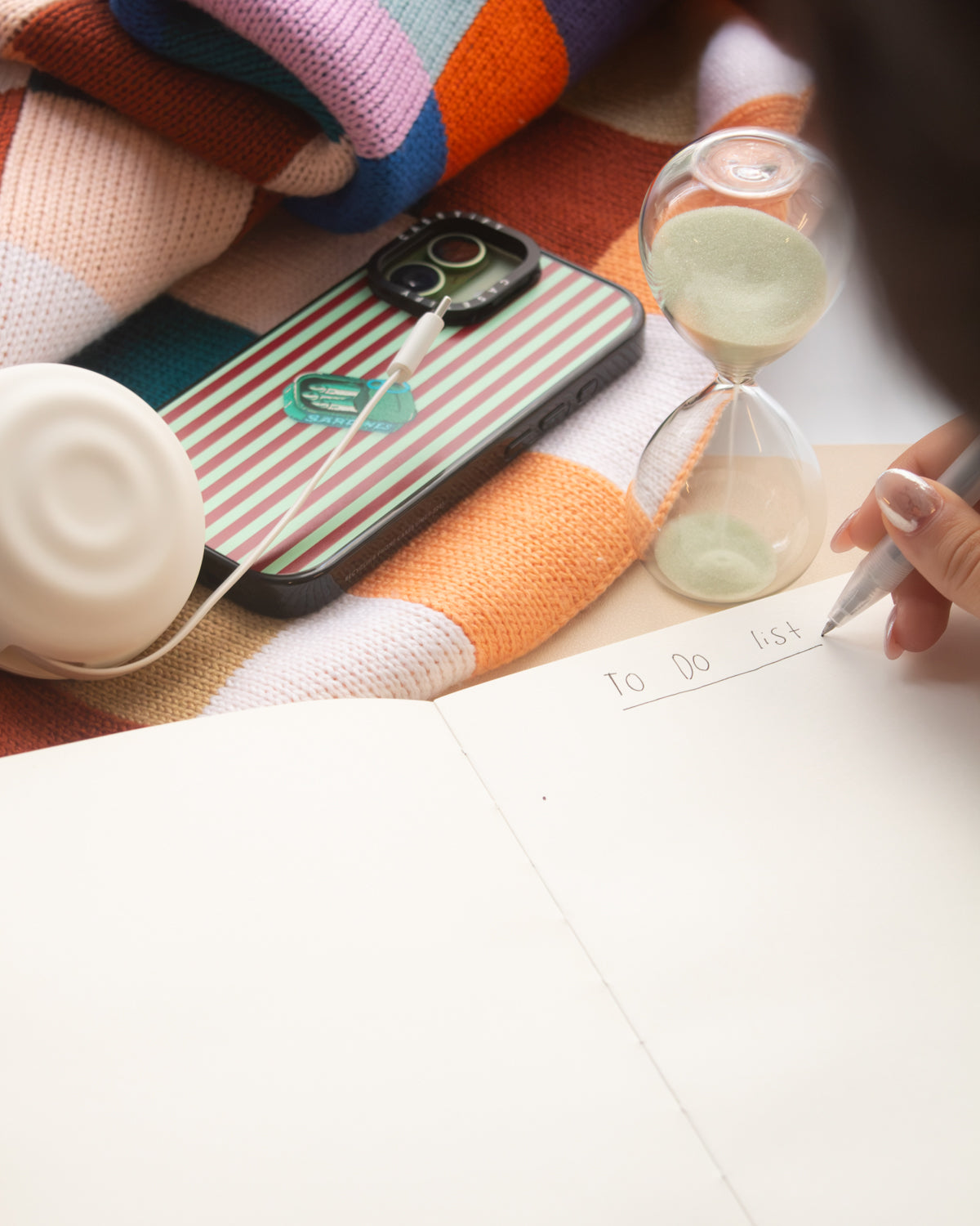 A close-up lifestyle shot of a hand writing on a white to-do list notebook page, surrounded by cozy props including a striped phone case, a glass hourglass, a white speaker, and a colorful knit blanket.