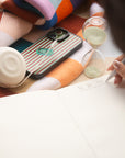 A close-up lifestyle shot of a hand writing on a white to-do list notebook page, surrounded by cozy props including a striped phone case, a glass hourglass, a white speaker, and a colorful knit blanket.
