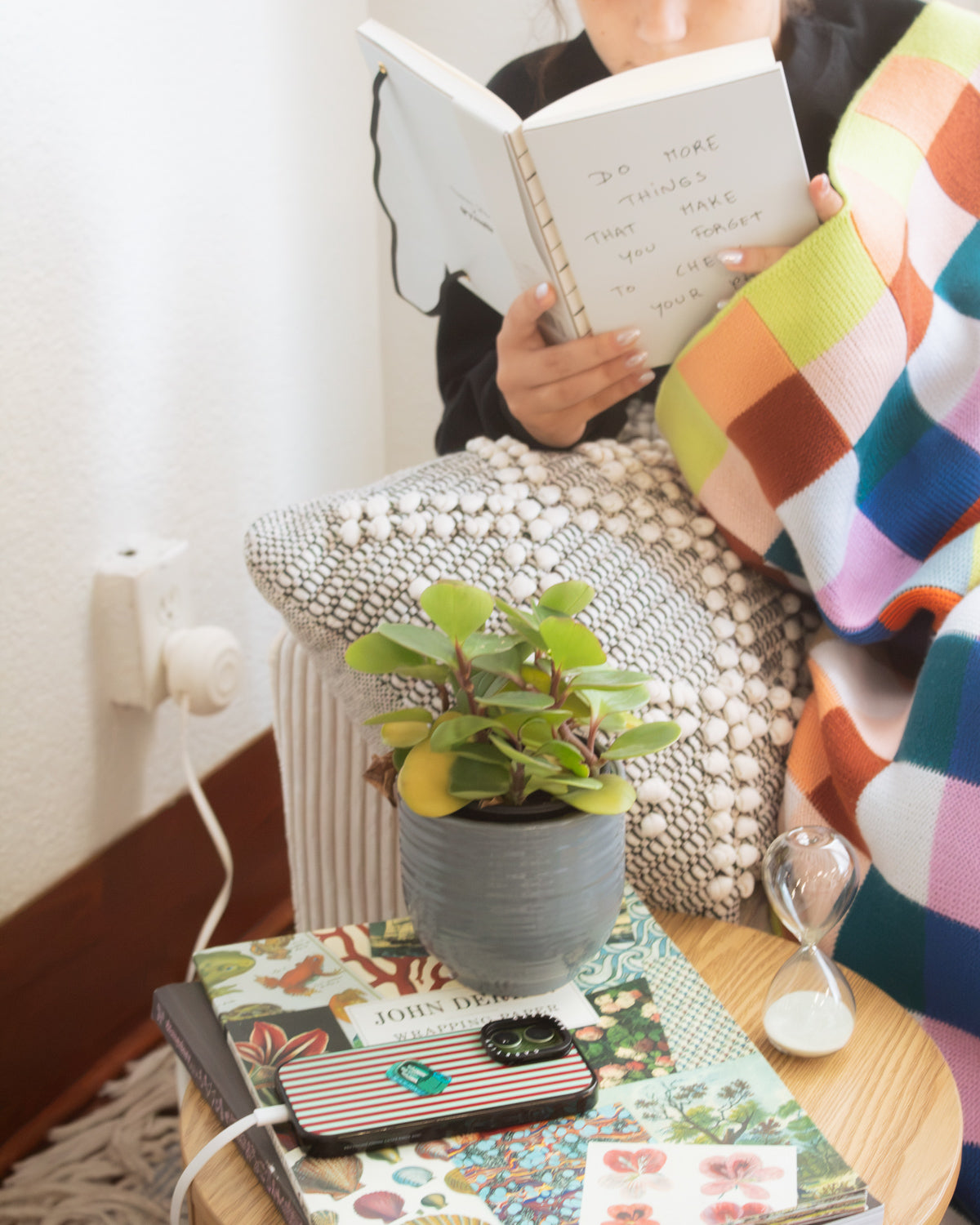 A cozy lifestyle scene showing a person wrapped in a colorful plaid blanket and reading a notebook on a wooden chair. In the foreground, a small potted succulent sits on a wooden table next to a striped phone case and a small hourglass.