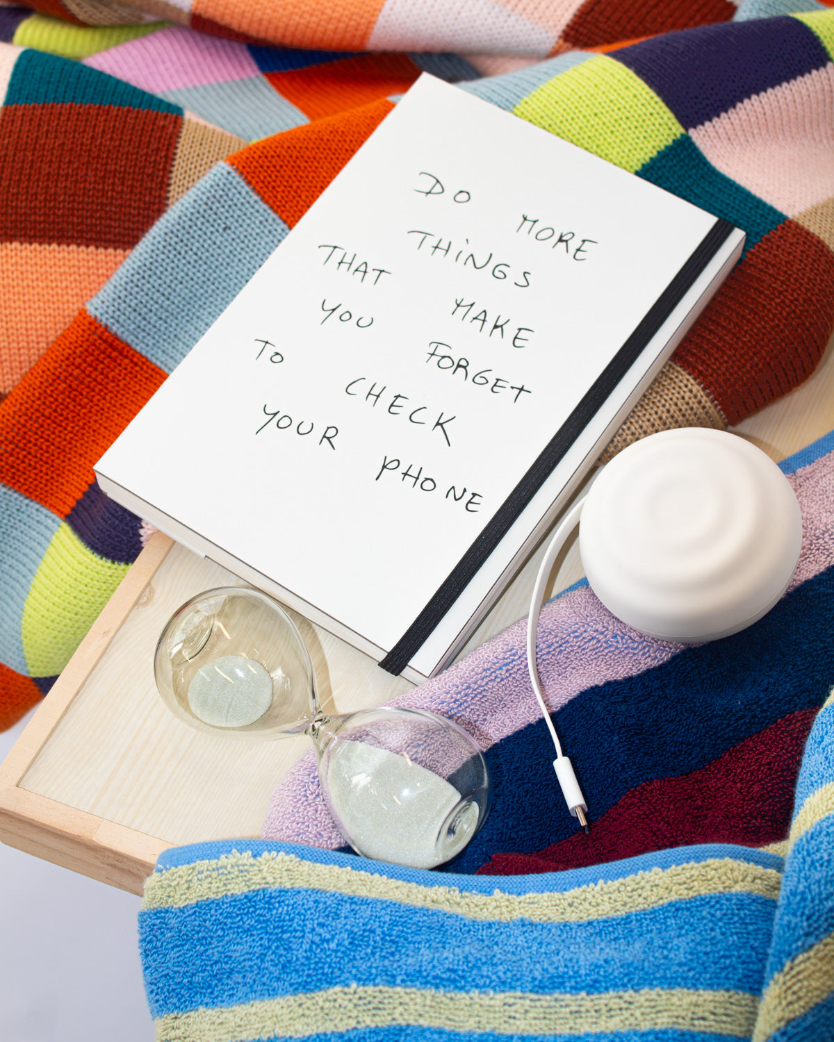 A white notebook with handwritten text reading 'Do more things that make you forget to check your phone' lies on a colorful striped blanket next to a white earbud case, a clear glass hourglass, and a white charging cable. The Dorm Move-in Set by The Mom Edit.