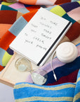 A white notebook with handwritten text reading 'Do more things that make you forget to check your phone' lies on a colorful striped blanket next to a white earbud case, a clear glass hourglass, and a white charging cable. The Dorm Move-in Set by The Mom Edit.