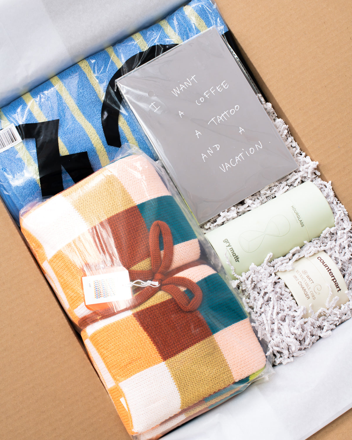 A curated gift box set for college students containing a folded plaid blanket in orange, teal, brown, and cream, a gray card that reads 'I WANT A COFFEE A TATTOO A VACATION AND A VACATION', a blue striped towel, and two green and white bottles nestled in white paper shred inside a brown cardboard box.