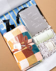 A curated gift box set for college students containing a folded plaid blanket in orange, teal, brown, and cream, a gray card that reads 'I WANT A COFFEE A TATTOO A VACATION AND A VACATION', a blue striped towel, and two green and white bottles nestled in white paper shred inside a brown cardboard box.