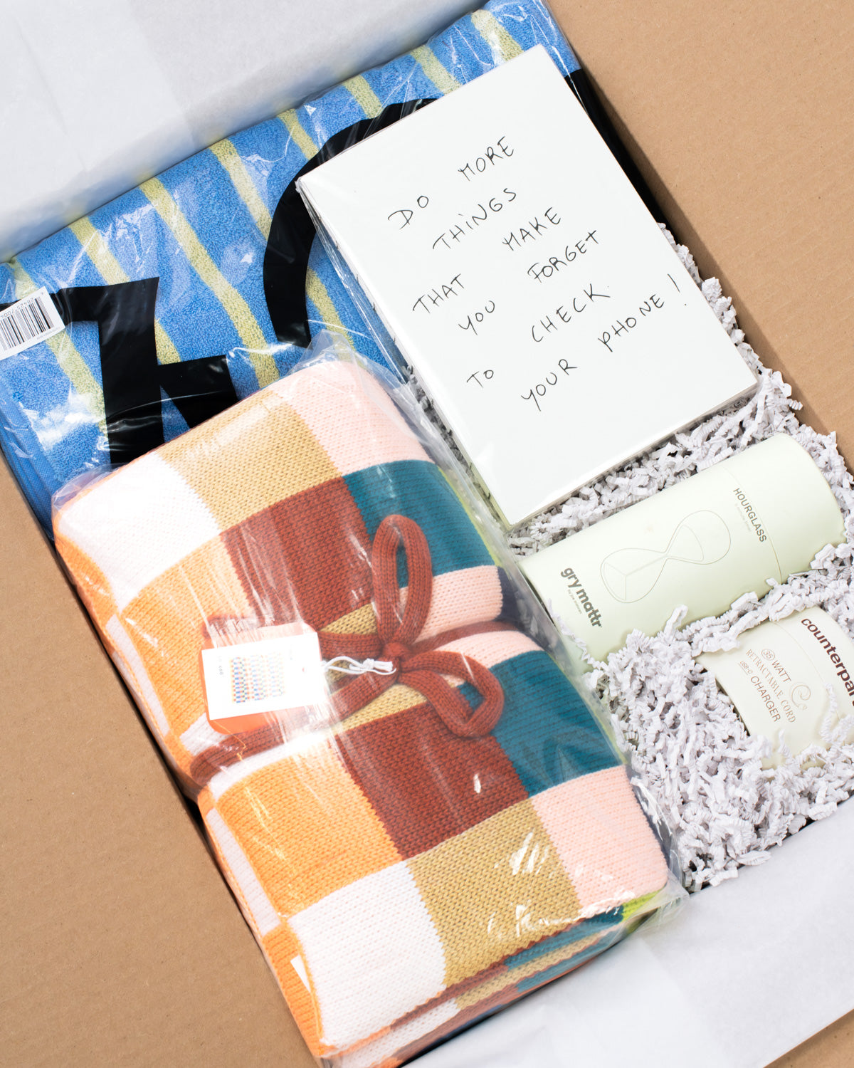 A curated gift box for college students containing a folded multicolor plaid blanket tied with a brown ribbon, a blue and yellow striped towel, a handwritten note, and two cream-colored bottles nestled in white crinkle paper inside a cardboard box.