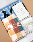 A curated gift box for college students containing a folded multicolor plaid blanket tied with a brown ribbon, a blue and yellow striped towel, a handwritten note, and two cream-colored bottles nestled in white crinkle paper inside a cardboard box.