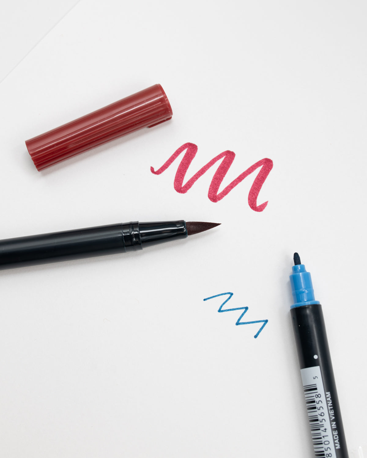 Red Tombow brush pen and blue fine-tip pen with test strokes on white paper.