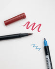 Red Tombow brush pen and blue fine-tip pen with test strokes on white paper.