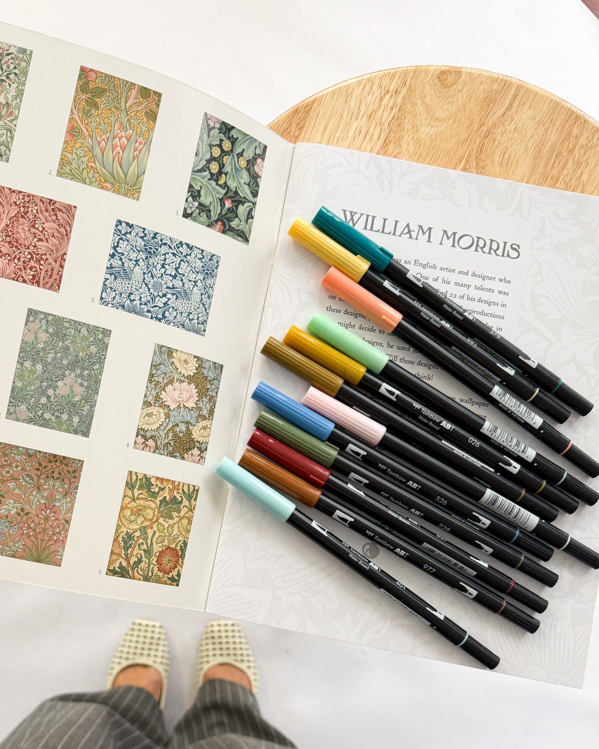 Tombow dual brush pens in muted greens, blues and ochres fanned over an open William Morris pattern book.
