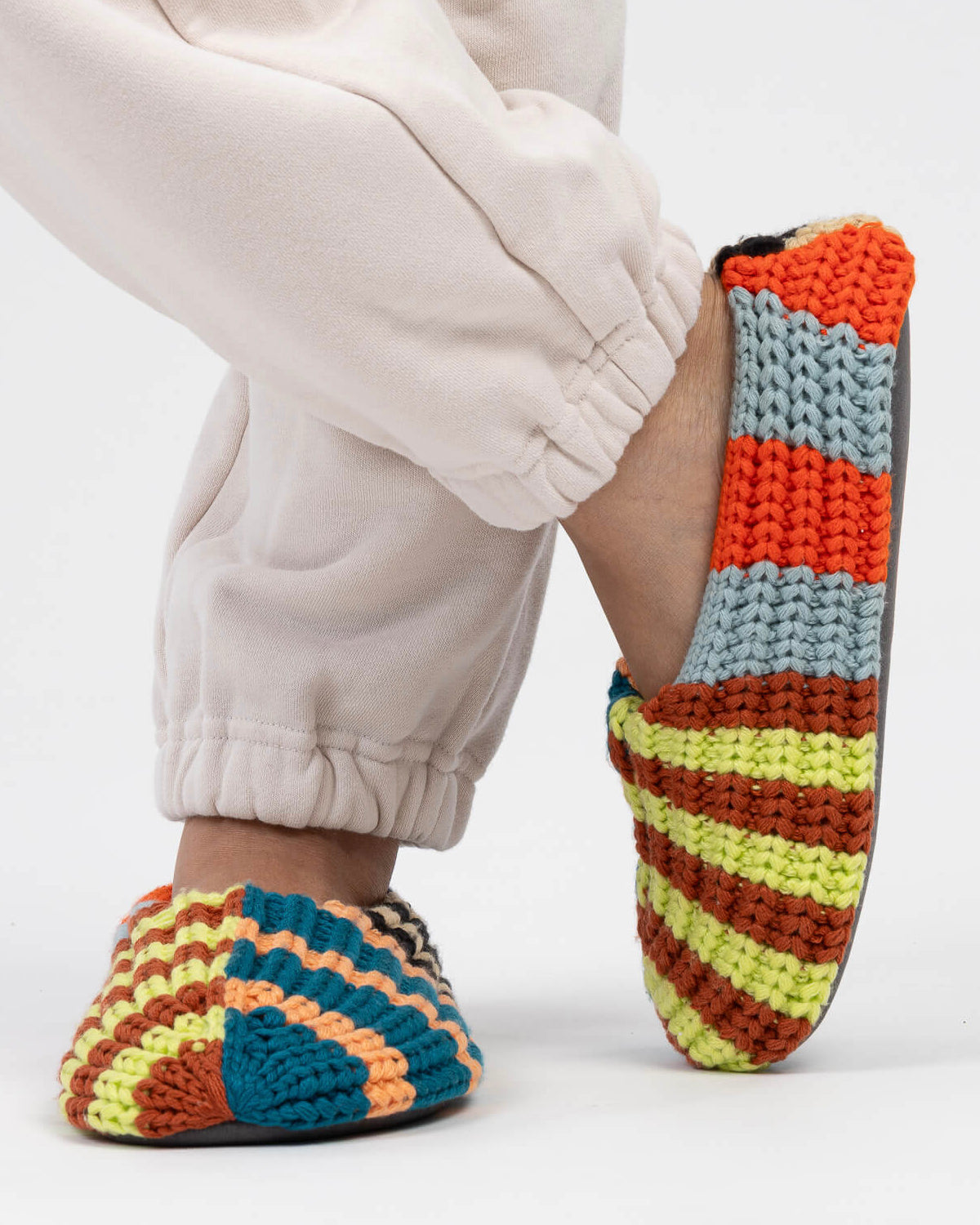 Person in cream joggers wearing Verloop knit house slippers in bold multicolor stripes
