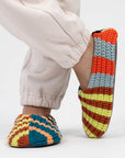 Person in cream joggers wearing Verloop knit house slippers in bold multicolor stripes