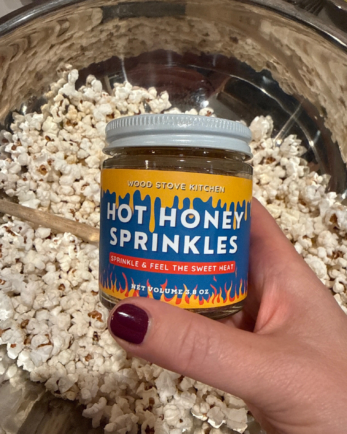 Hand holding a jar of Wood Stove Kitchen Hot Honey Sprinkles over a bowl of popcorn