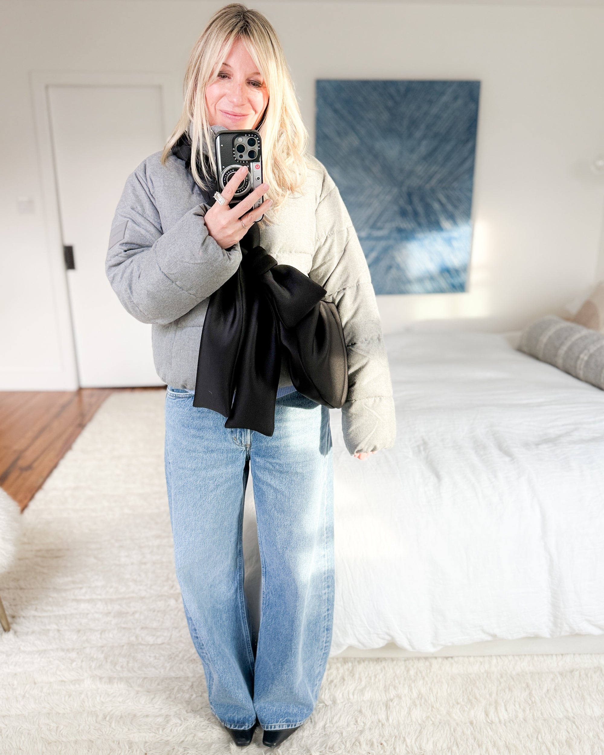 Woman styling a black bow crossbody bag over a gray puffer jacket and wide-leg jeans.